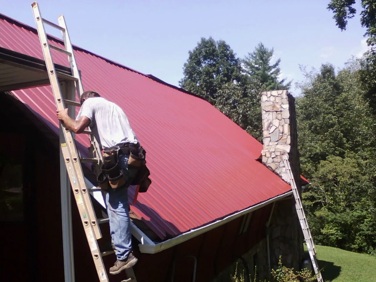 Roofer on a ladder beside a red metal roof and stone chimney — Soffit & Fascia Repair by Roofing Brothers in Pottstown